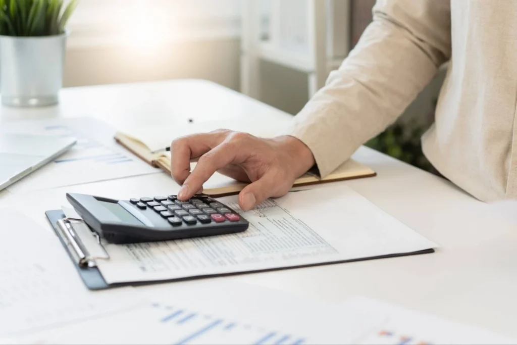 view of woman's arm using calculator on desk covered in financial documents so she can calculate savings goals