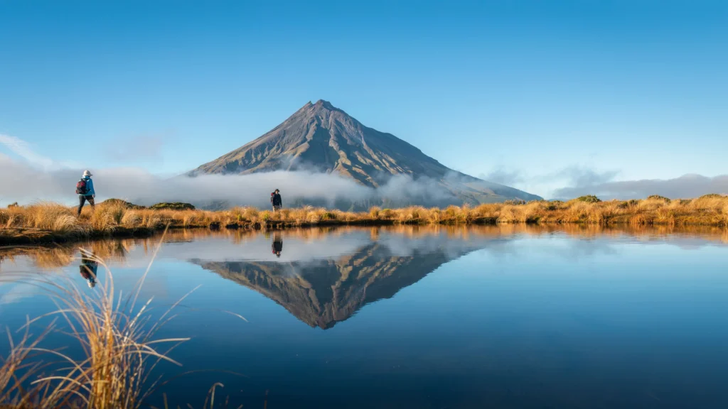 Two hikers on the Pouakai Crossing trail along a reflective lake with Mount Taranaki and clouds in background.