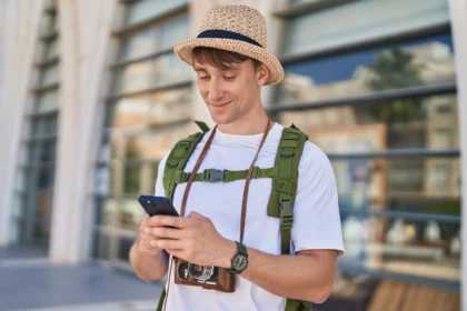 Young man wearing a straw hat and backpack, looking at his smartphone outdoors near modern glass buildings.