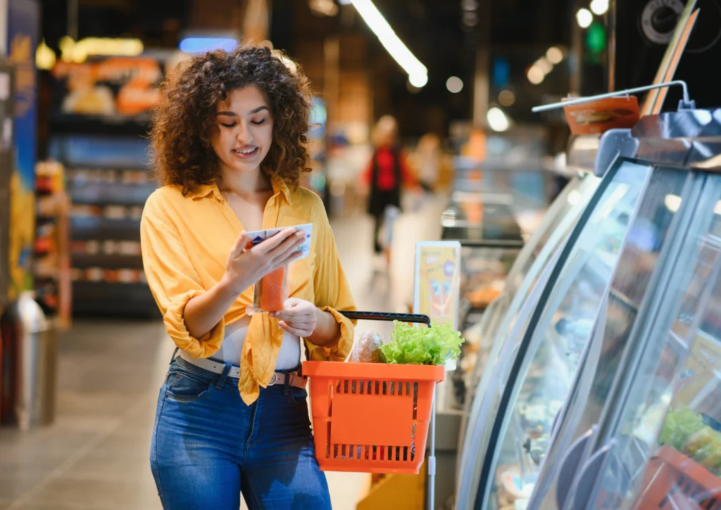 A woman in a yellow shirt smiles while holding a smartphone and a basket of groceries in a store aisle, giving a relaxed, purposeful feel.