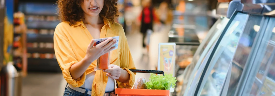 A woman in a yellow shirt smiles while holding a smartphone and a basket of groceries in a store aisle, giving a relaxed, purposeful feel.