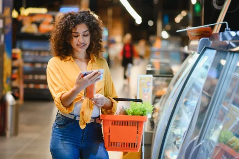 A woman in a yellow shirt smiles while holding a smartphone and a basket of groceries in a store aisle, giving a relaxed, purposeful feel.
