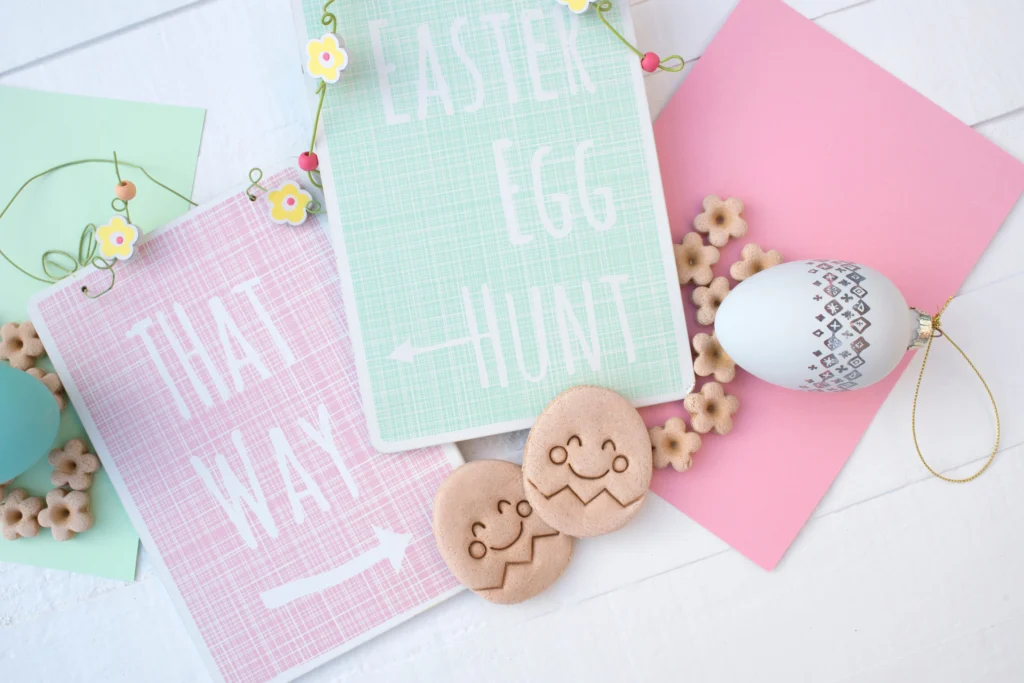 pieces of paper and Easter decorations laid out. Aqua piece of paper says "Easter Egg Hunt" and baby pink says "That Way" with an arrow