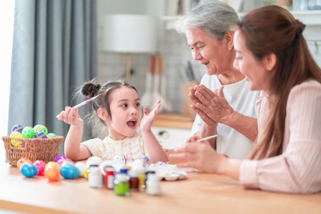 mother and grandma painting Easter eggs with a cute little girl at a wooden table, with different paint pot colours laid out on table