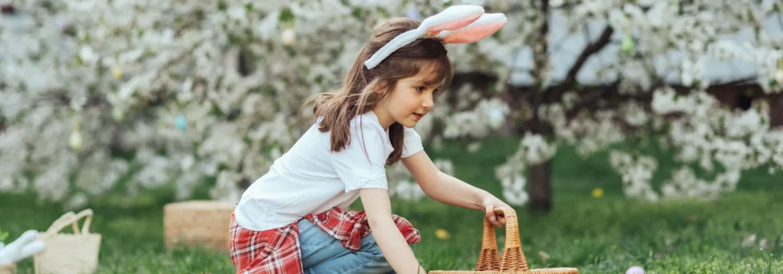 little girl with bunny ears holding a wicker basket picking up a green egg in the grass doing an Easter egg hunt