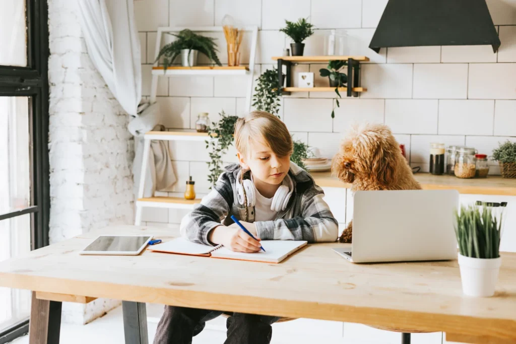 Kid writing in workbook with laptop in front of him as he gets taught online by tutor