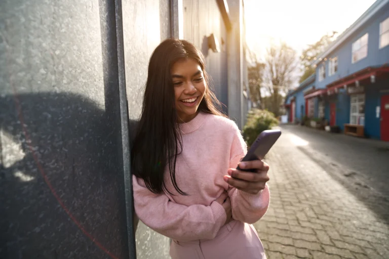 Woman chilling outside on the pathway between buildings happily looking at her phone as she applies for a secured personal loan
