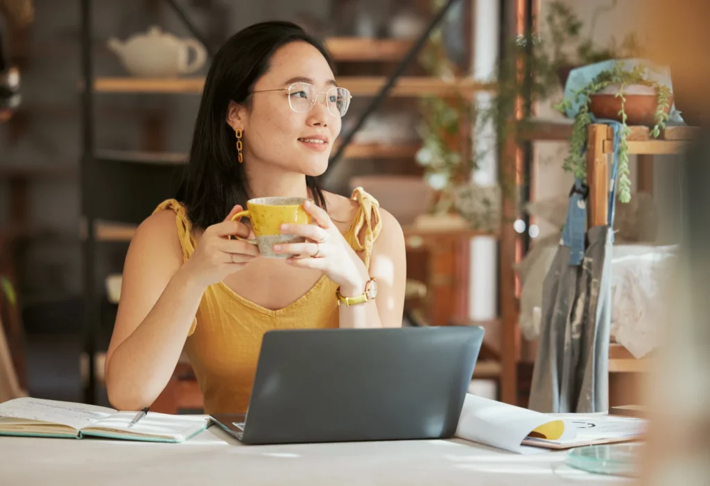 Woman sitting at a desk with laptop holding a mug, taking a break from writing an eBook to make extra money online